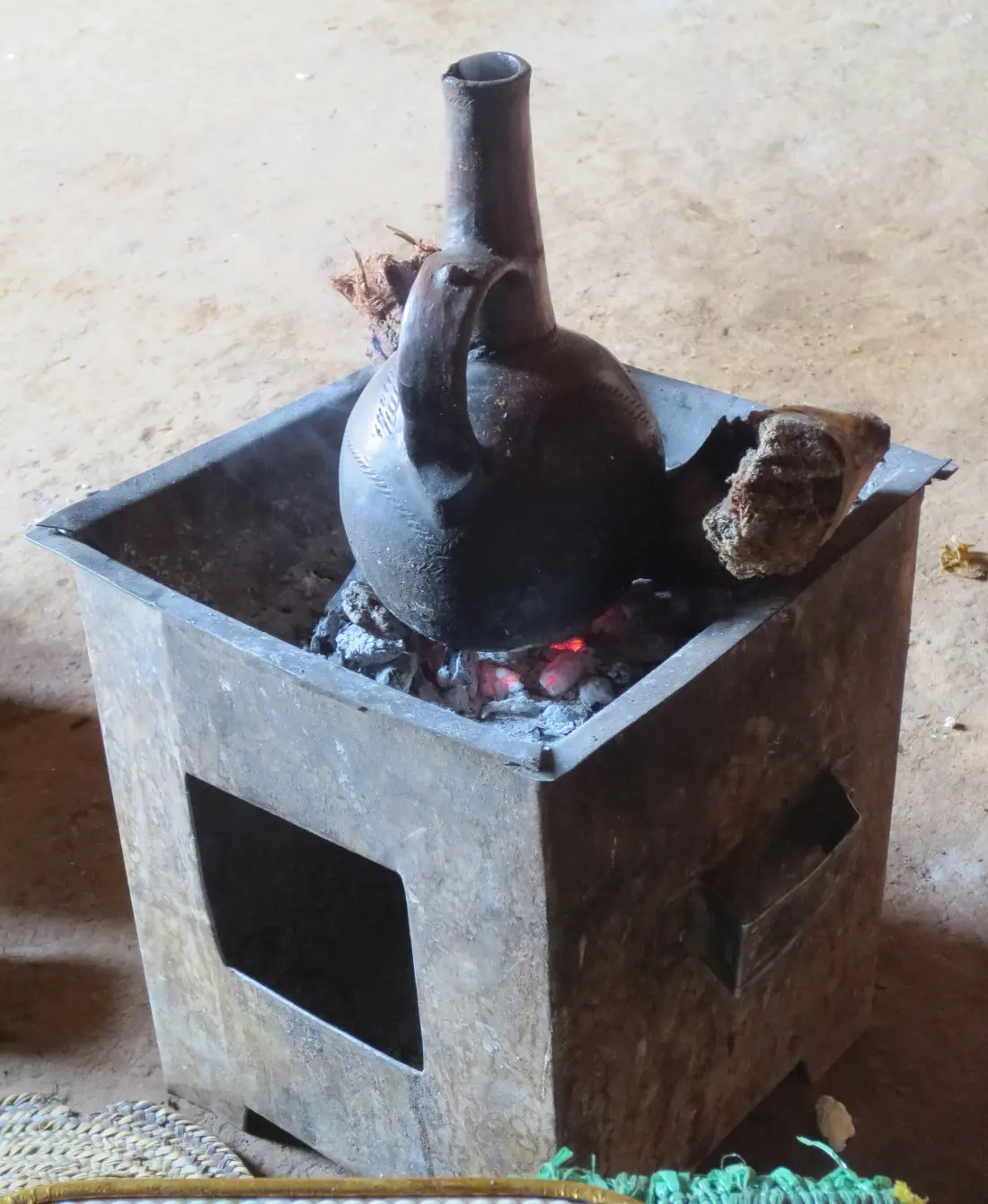 A black clay jebena coffee pot sitting over glowing coals in a metal stove
